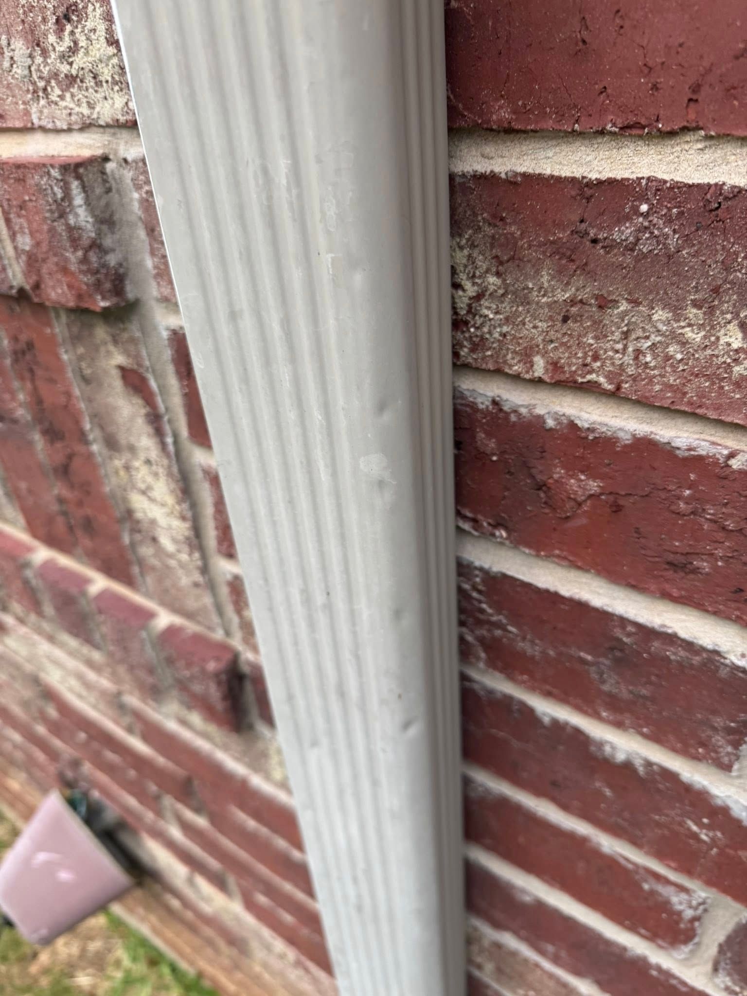 Close-up of a white downspout mounted on a red brick wall near the ground.