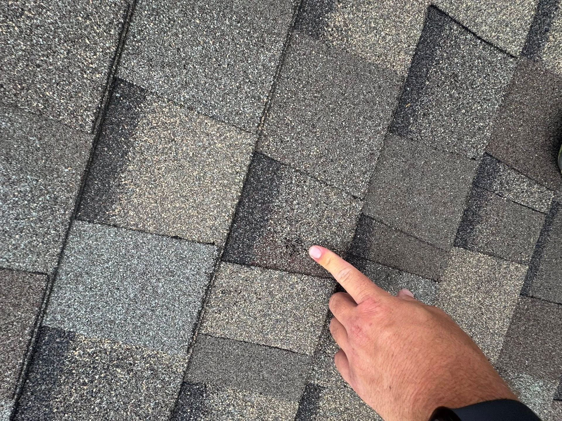 Hand pointing at gray roof shingles with visible wear and discoloration