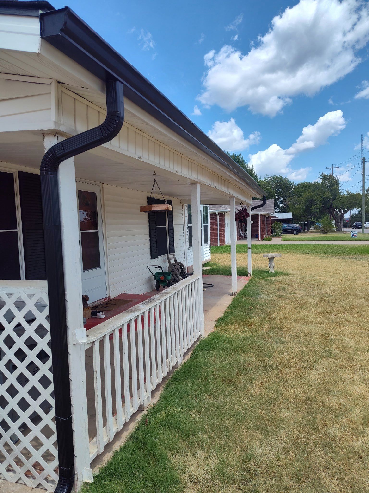 White house porch with lattice railing, black gutters, and grassy yard under a blue sky.