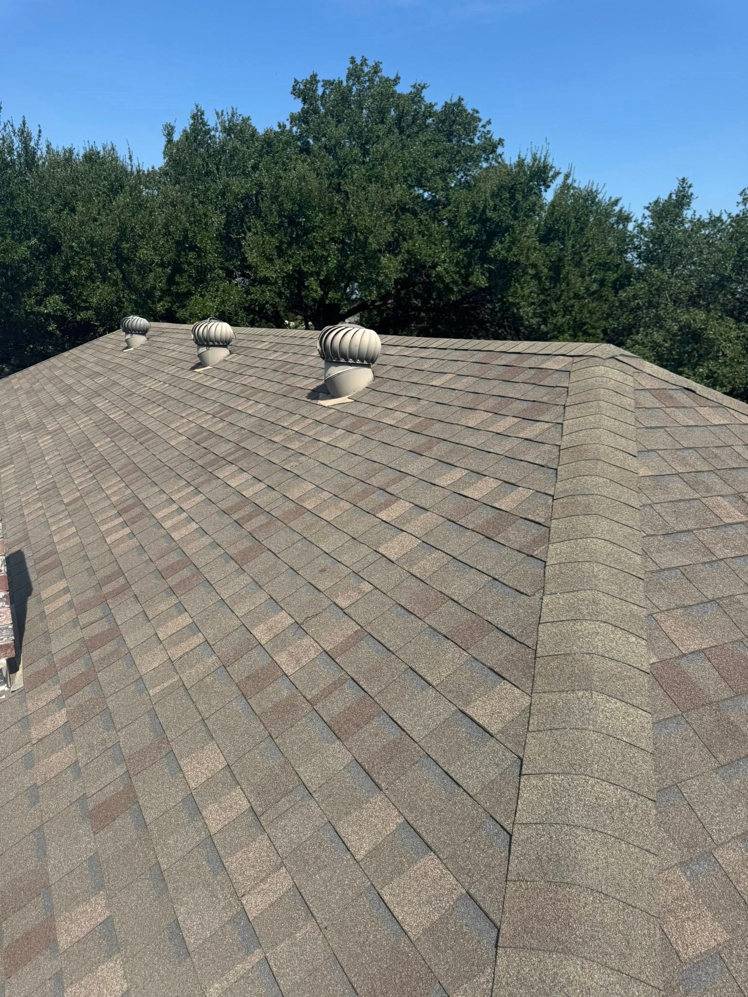 Weathered shingle roof with a vent pipe and trees in the background under a clear blue sky