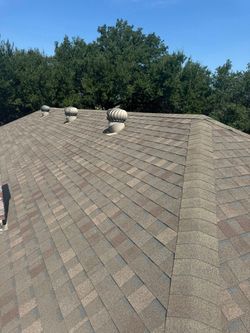 View from a gray shingled roof toward a stone chimney, green trees, and blue sky with clouds