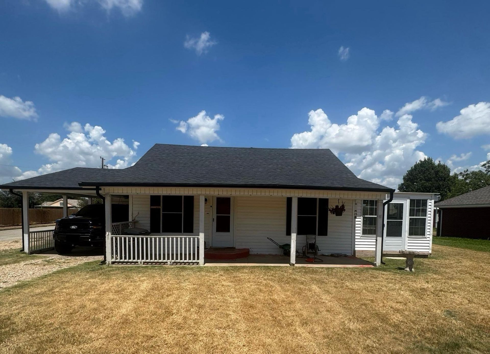 Single-story white house with black roof, front porch, and carport under a blue sky