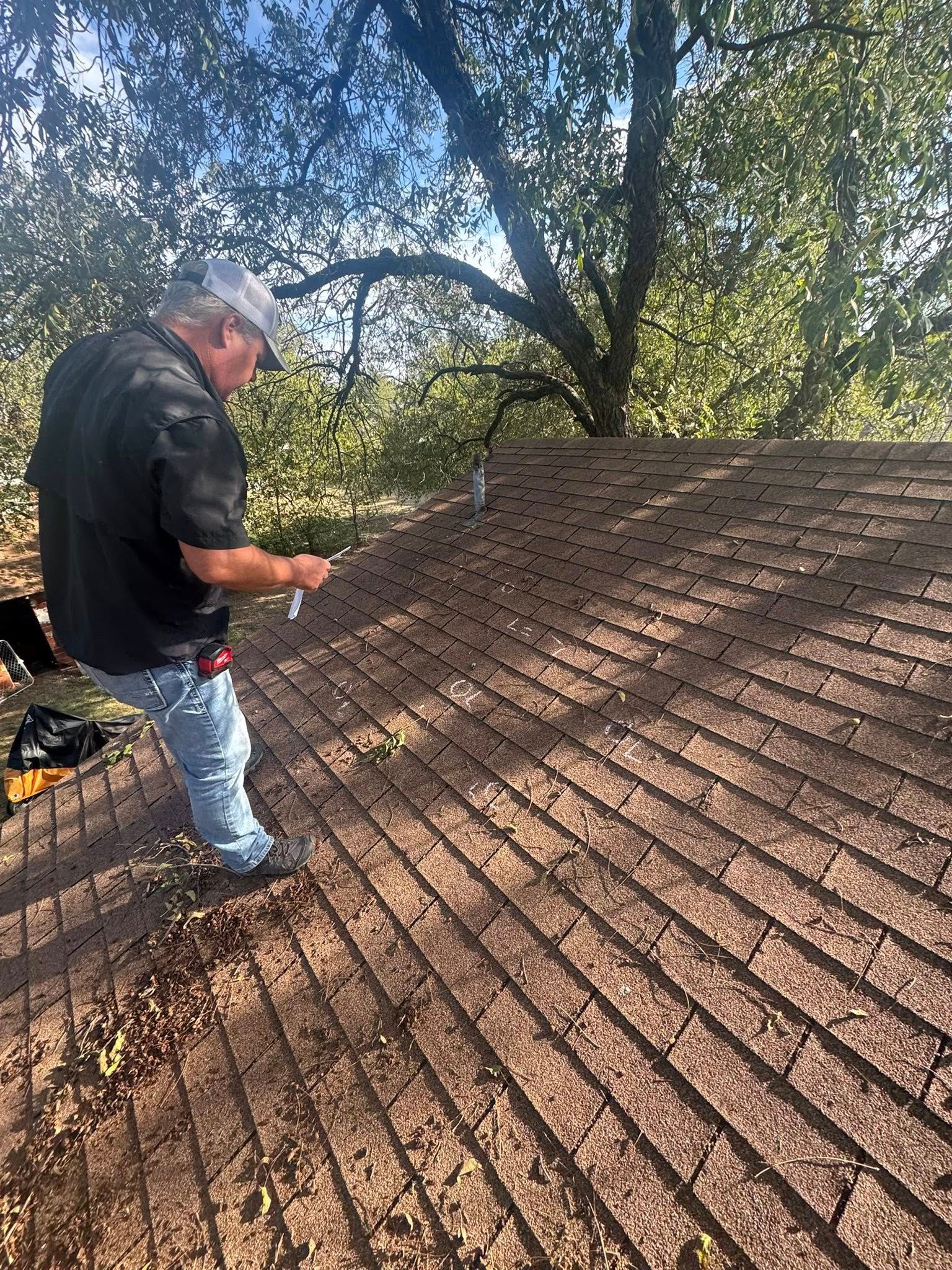 Worker inspecting a shingled roof beside trees in daylight