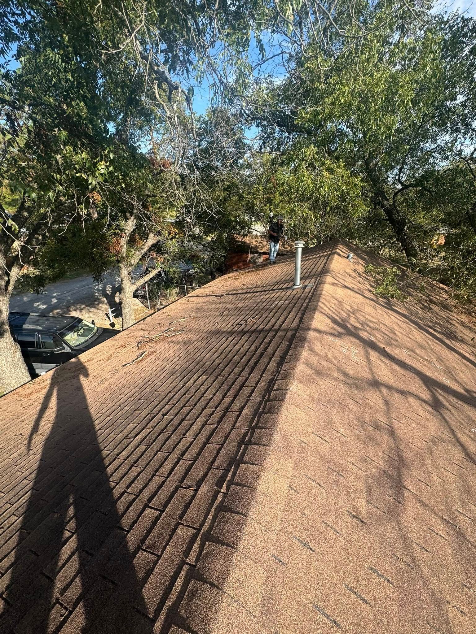 Rooftop with brown shingles and vent pipes, surrounded by leafy trees under a clear blue sky.