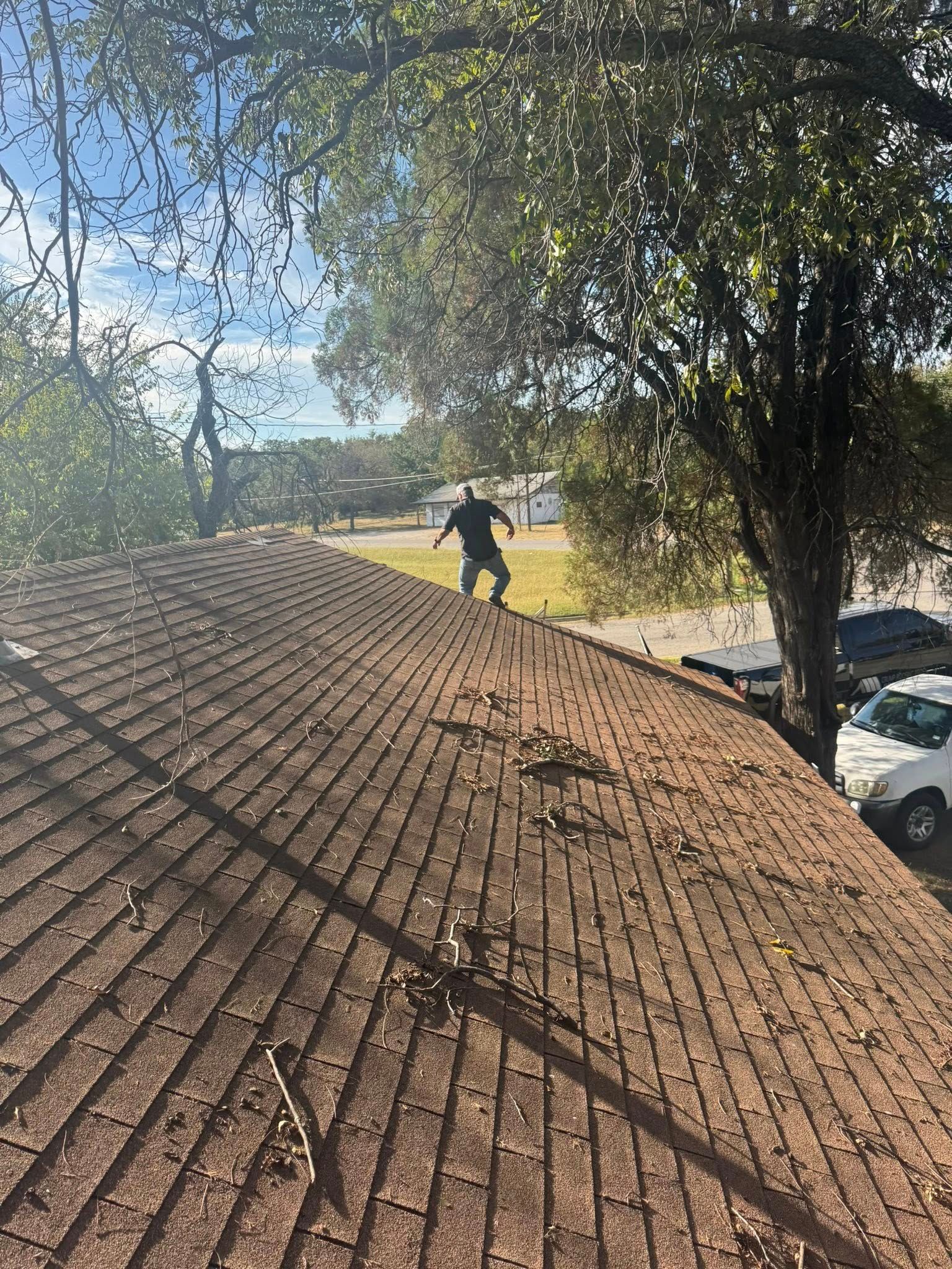 Worker on a shingled roof with a leaf blower, trees and yard in the background.