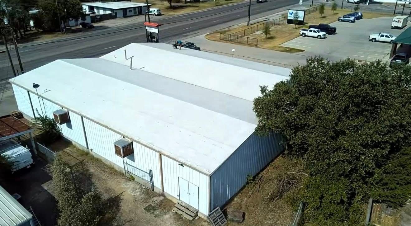 Aerial view of a large white warehouse with a blue side wall beside a road and parked cars.
