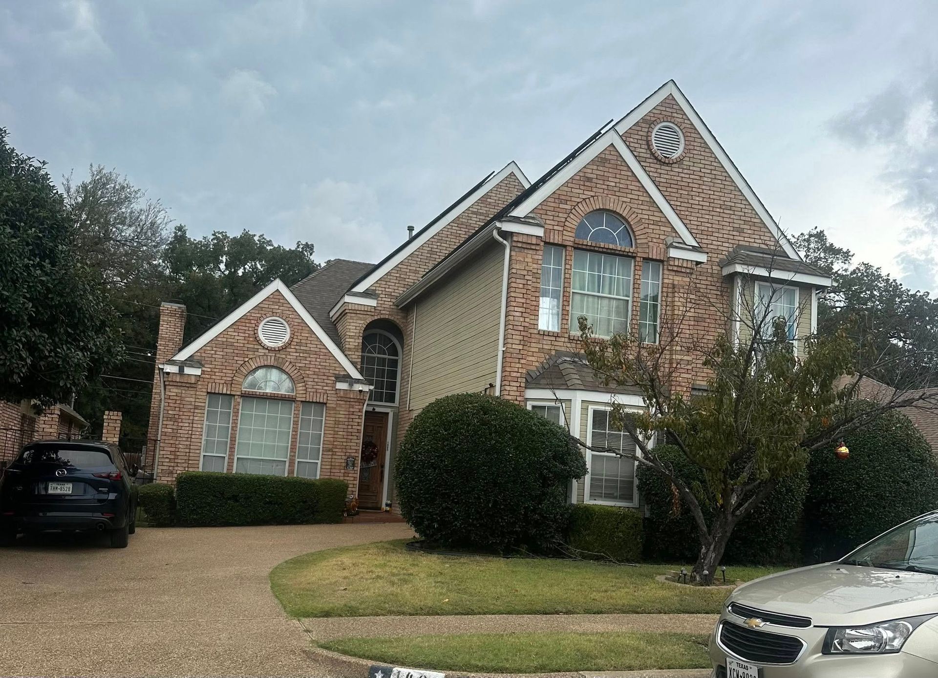 Brick suburban house with large windows, front lawn, and two parked cars under a cloudy sky