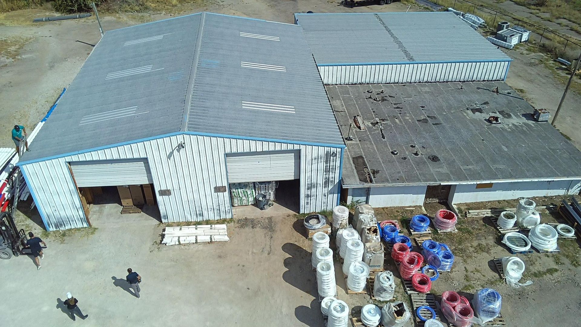 Aerial view of a large corrugated-roof warehouse with stacked pipes and drums outside.