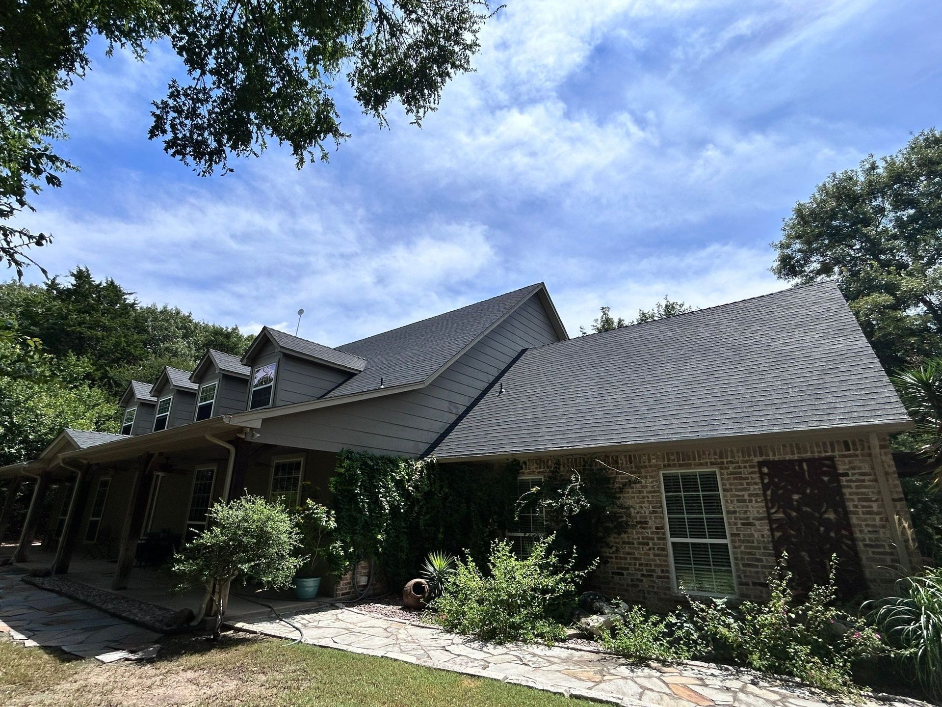 Single-story brick house with a wavy gray roof, surrounded by trees and greenery under a blue sky