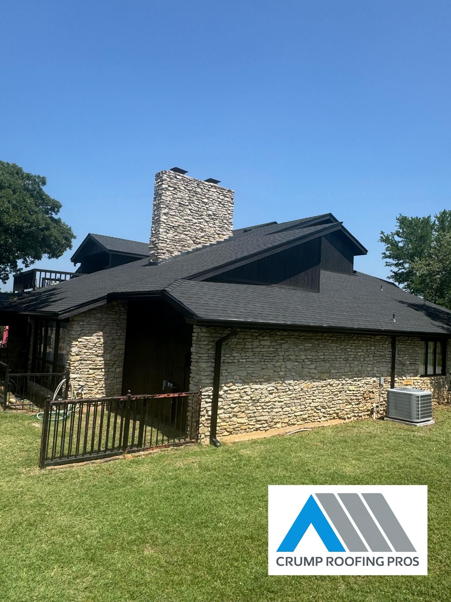 Stone house with dark shingled roof, chimney, and fenced yard under a clear blue sky