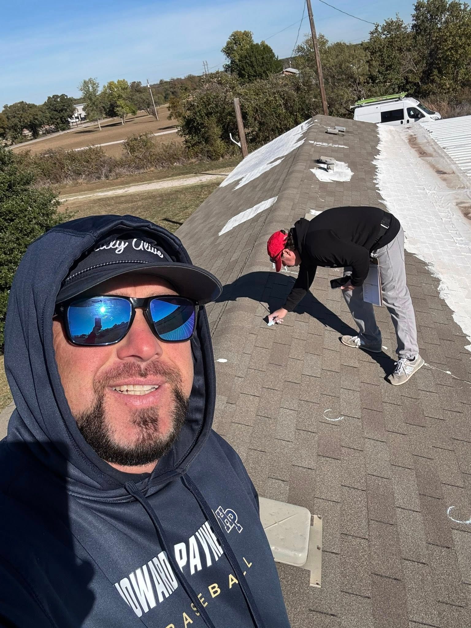 Man taking a selfie on a gravel road while two people work by a white barrier in the background