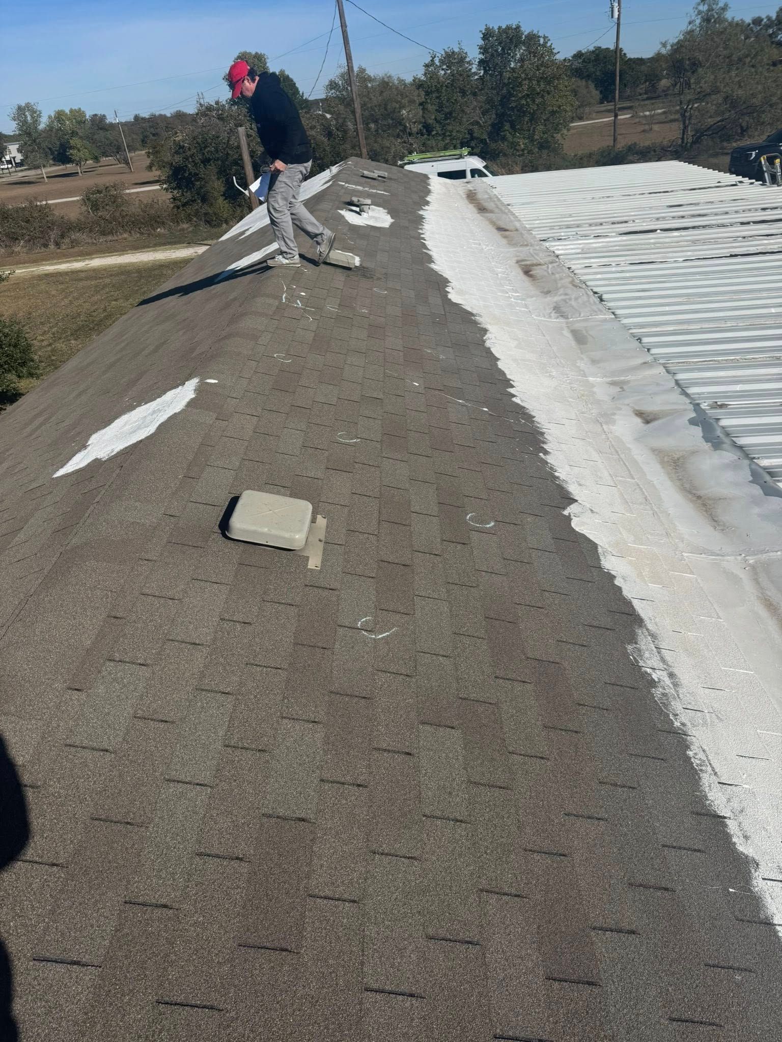 Worker on a sloped rooftop repairing shingles beside a white tarp-covered section.