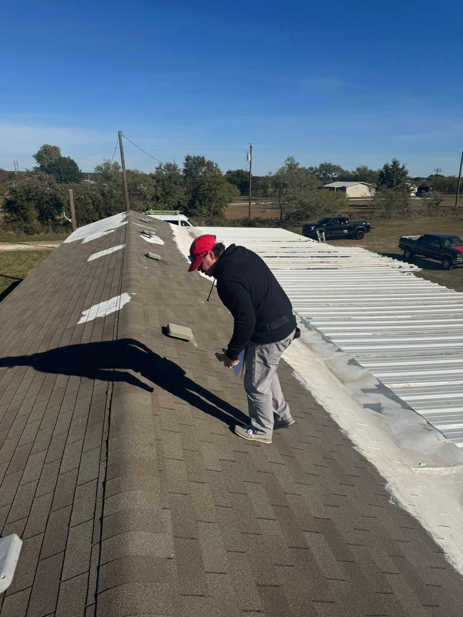 Worker on a rooftop inspecting a sloped gravel surface beside a white metal roof under a clear blue sky