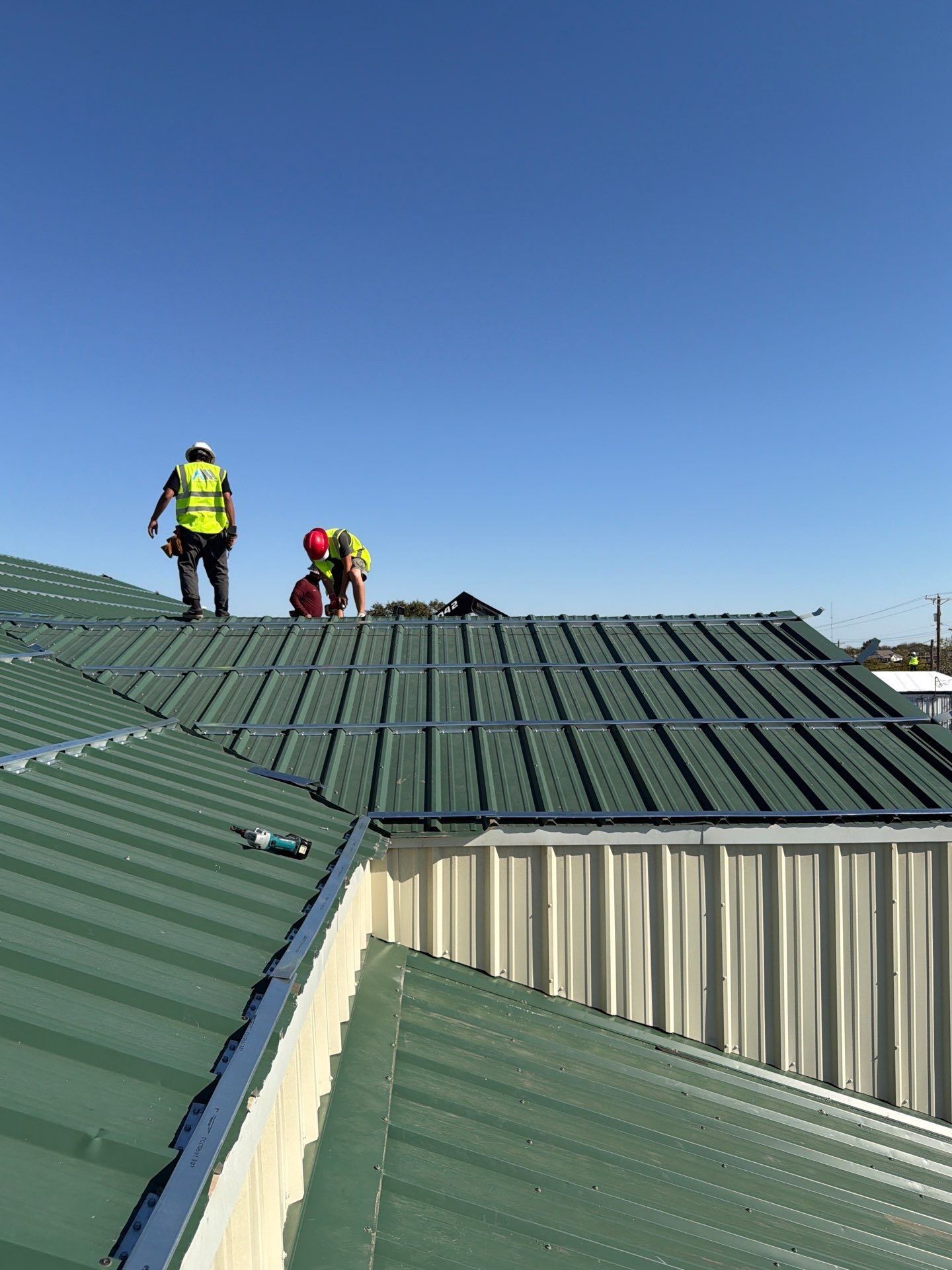 Workers on a green metal roof, inspecting or repairing it under a clear blue sky.