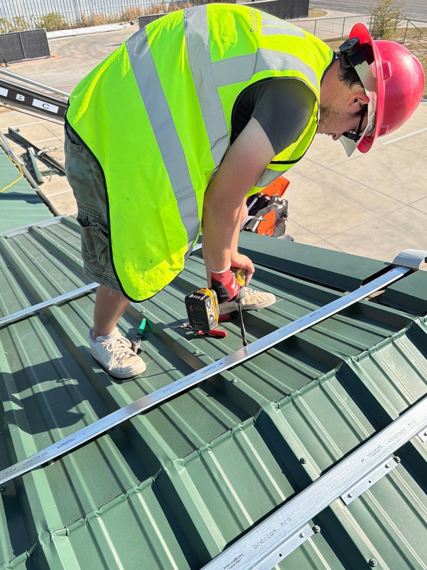 Worker in neon safety vest and red hard hat fastening metal panels on a rooftop.