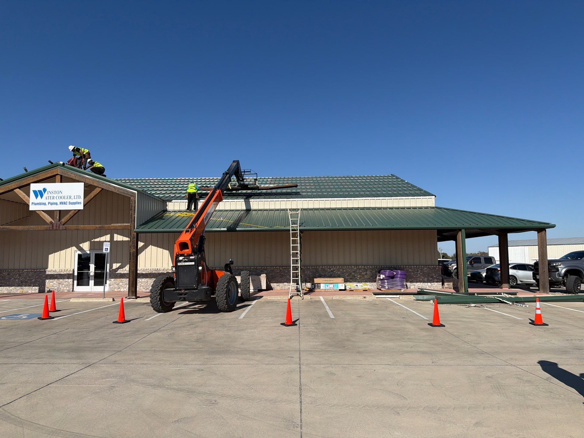 Construction lift outside a building with orange cones and a ladder under a green roof.