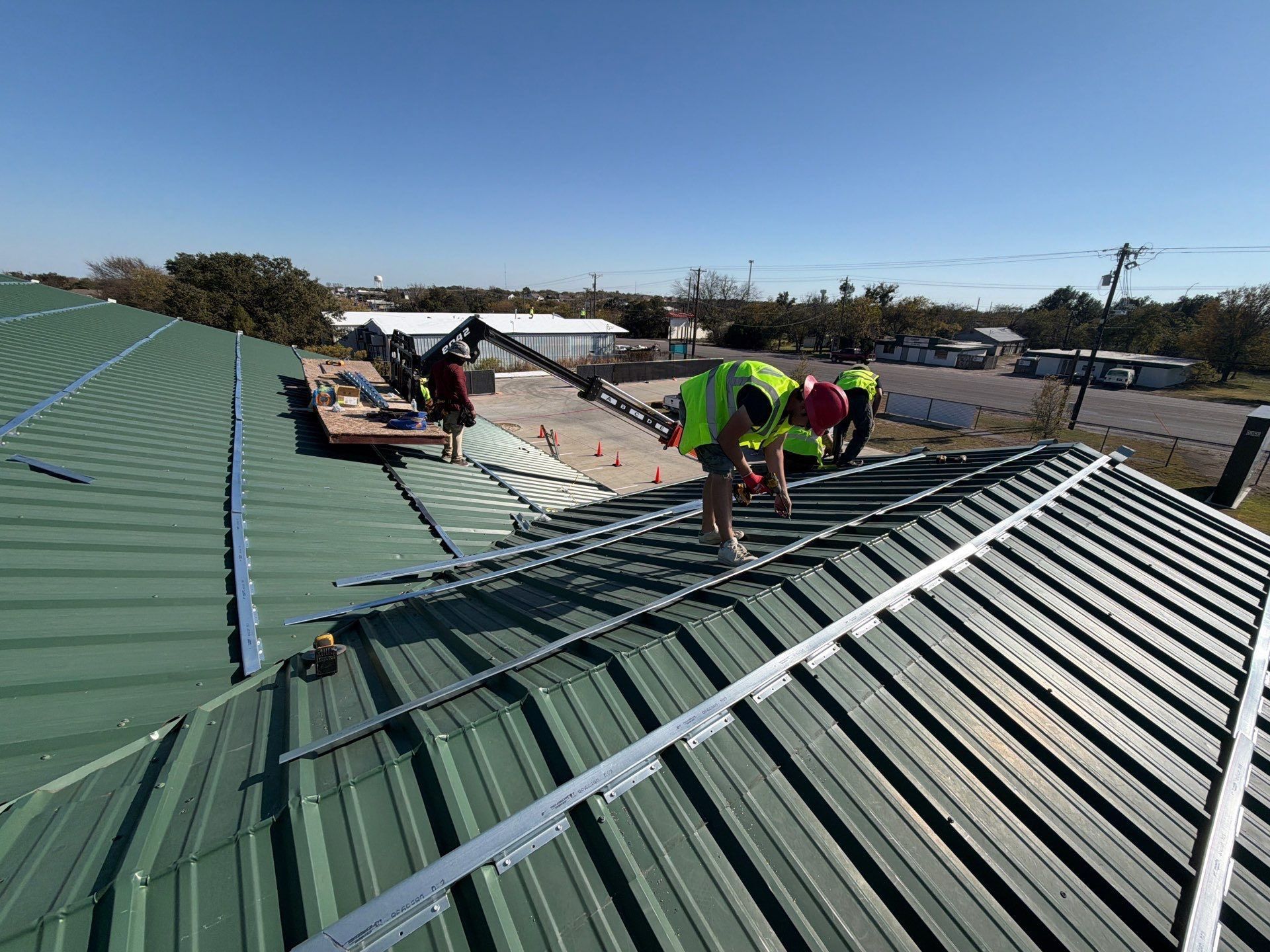 Workers installing green metal roofing on a sloped roof under a clear blue sky