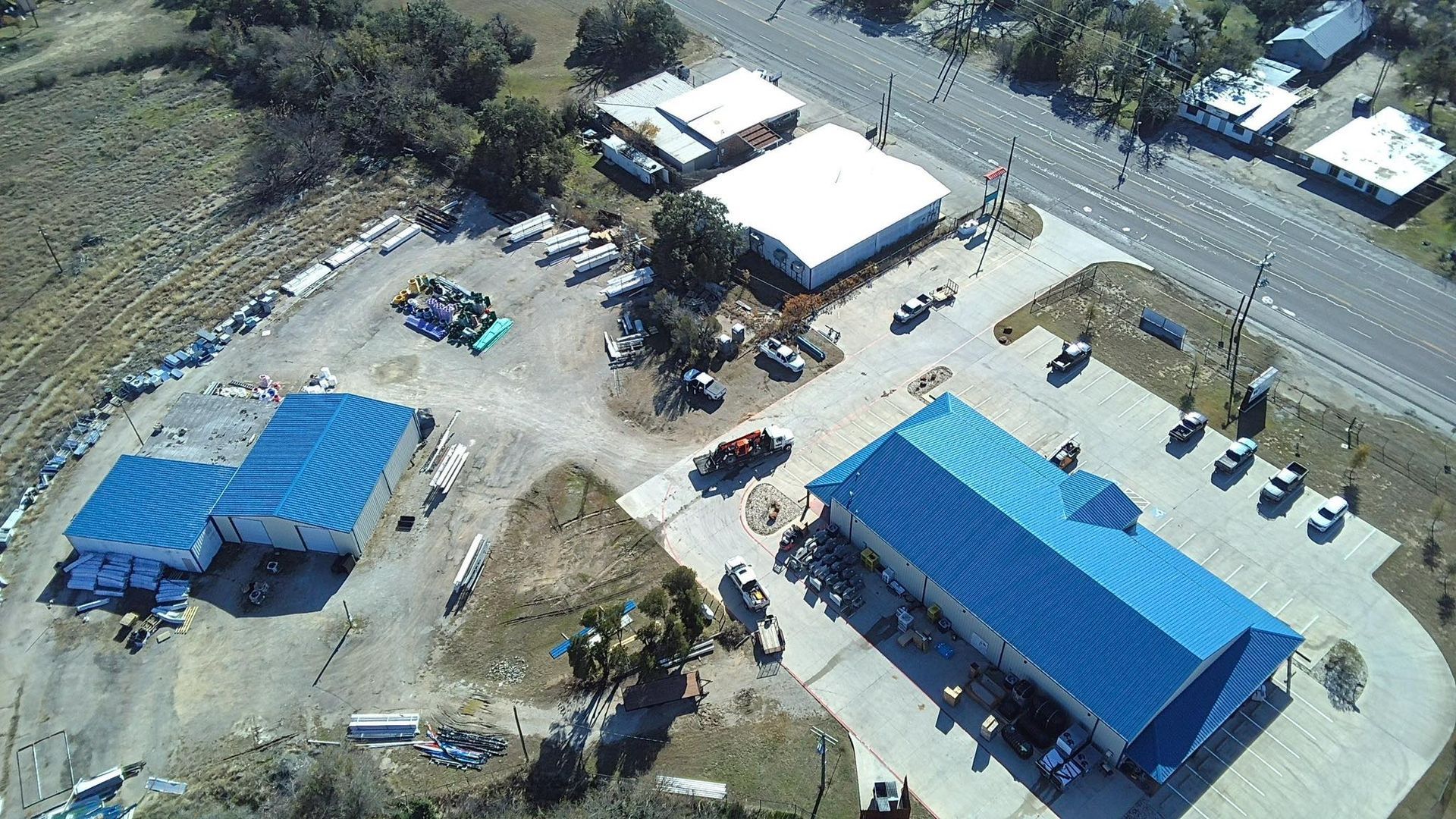 Aerial view of a rural facility with blue-roofed buildings, parked vehicles, and dirt lots beside a road.