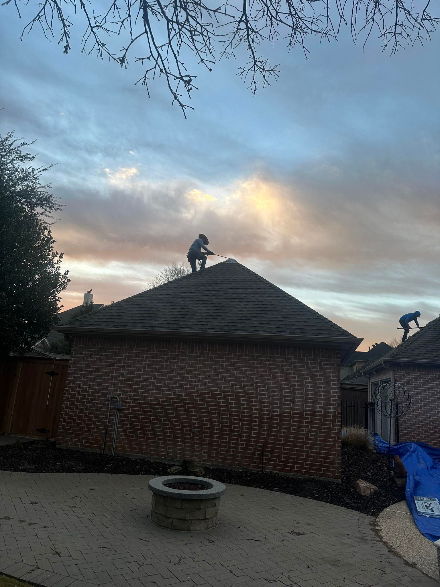 Workers on a thatched roof at dusk, with a brick courtyard and cloudy sky.