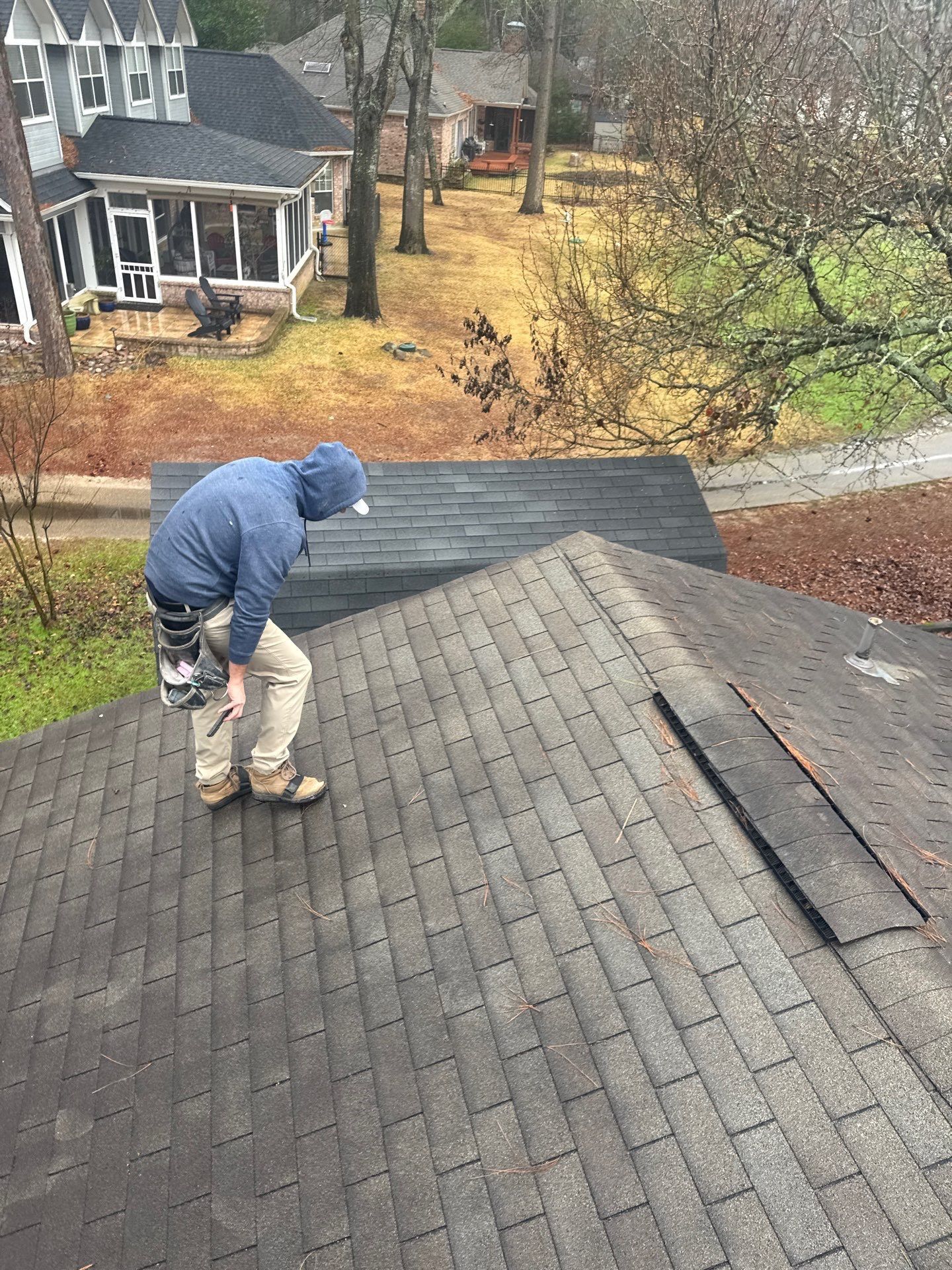 Worker in blue jacket repairs shingles on a steep residential roof, with houses and trees in the background.