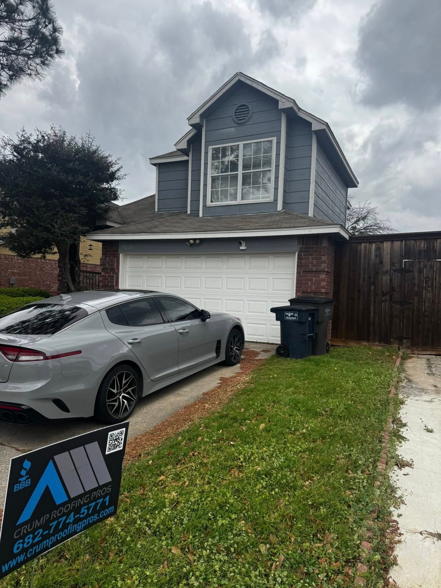 Gray house with white garage, parked silver car, and a yard sign on a wet driveway under cloudy skies