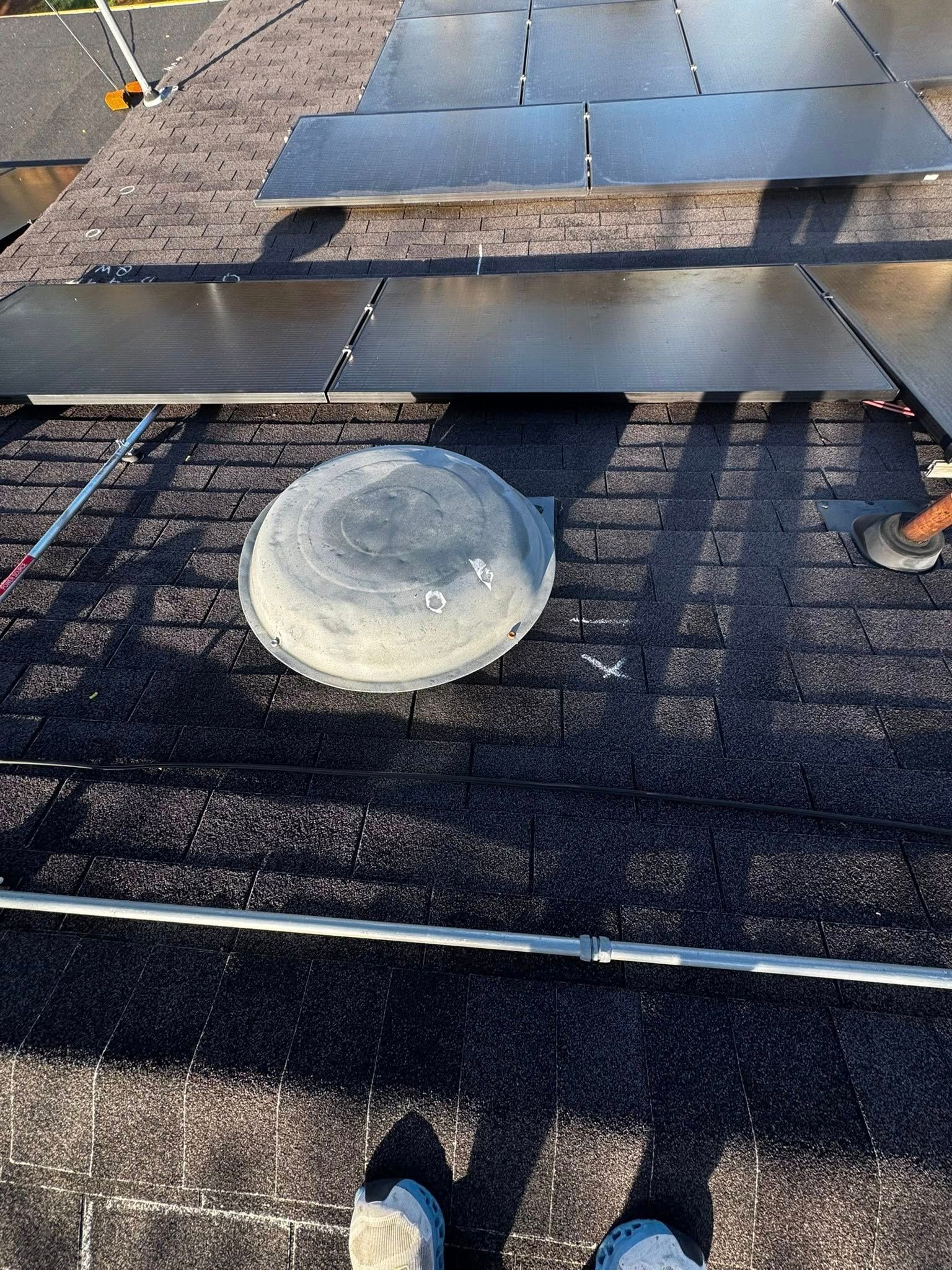 Roof under repair with shingles, a vent pipe, and a worker’s feet visible near the edge