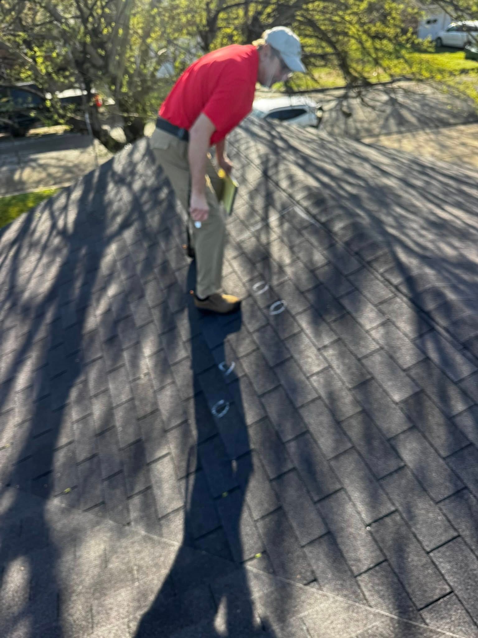 Worker in red shirt and tan pants inspecting a shingled roof under bright sunlight.