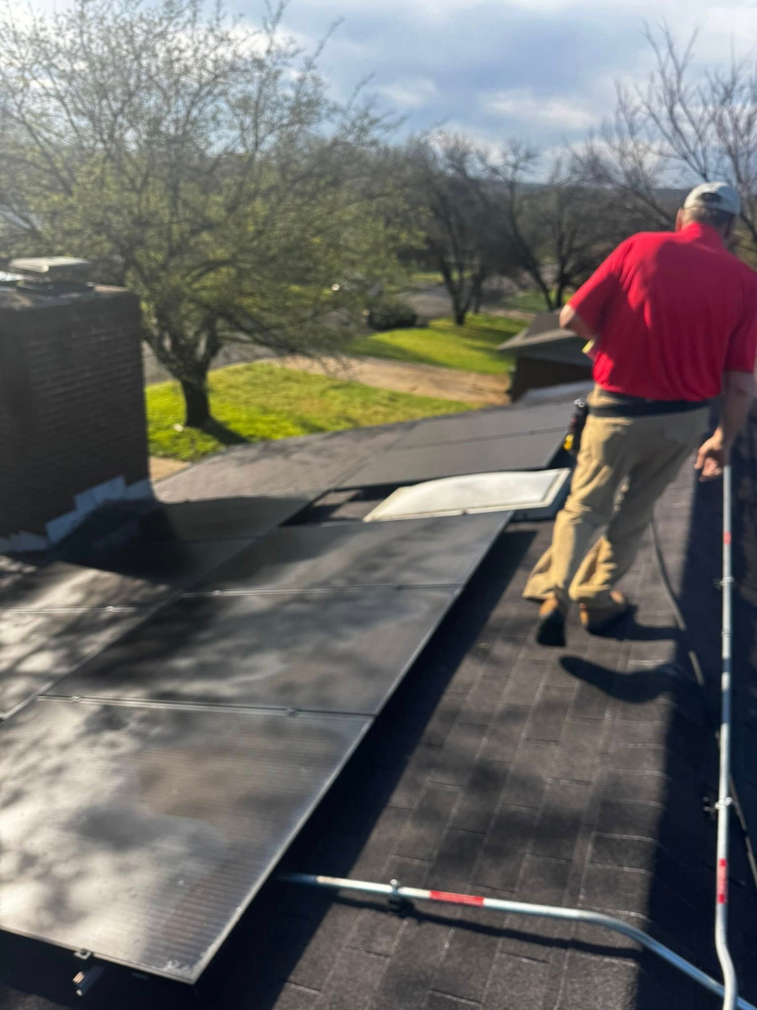 Worker in a red shirt inspecting a rooftop with black roofing material near a tree-lined area.