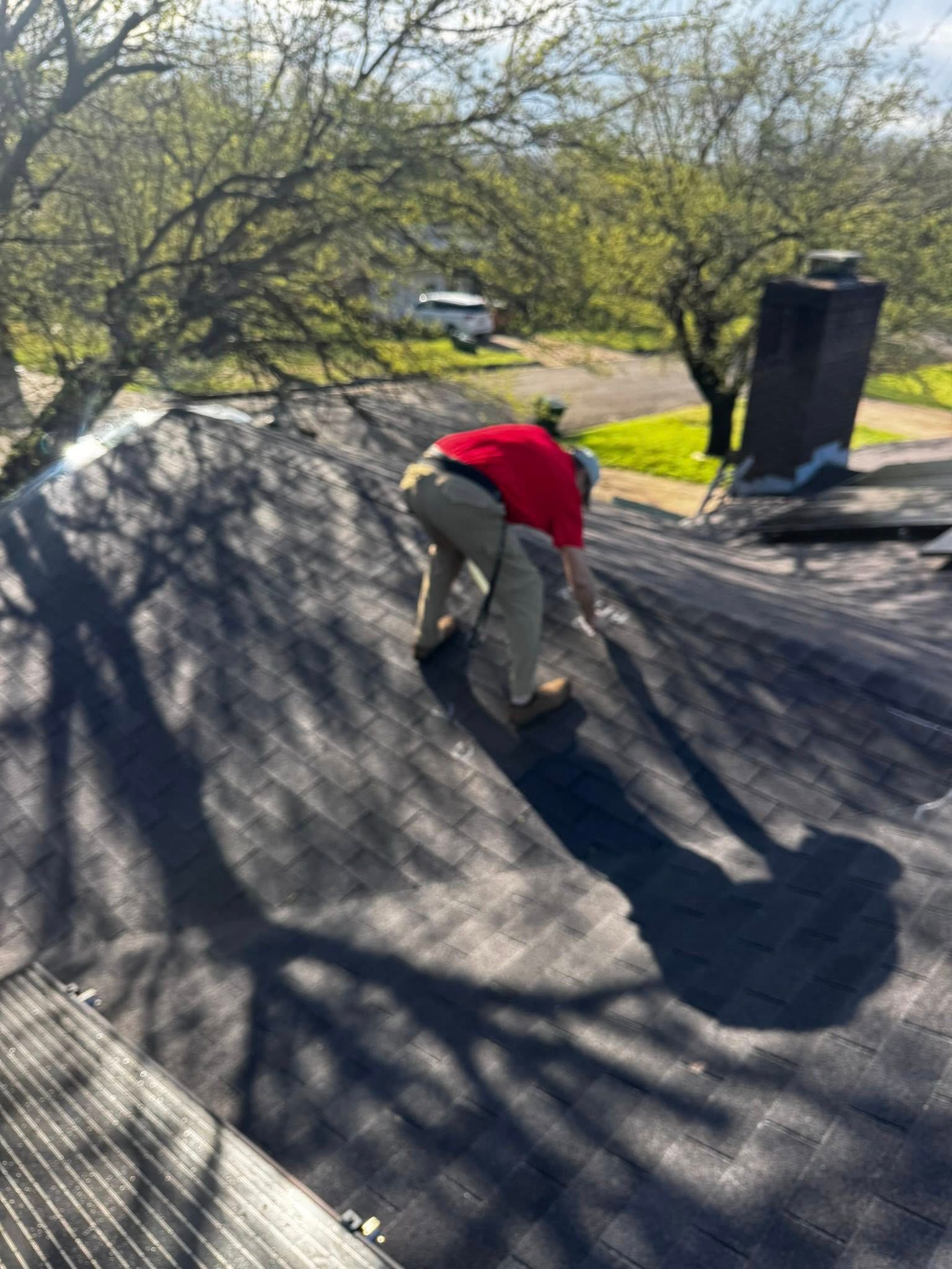 Person in a red jacket crouches on a shingled roof beside trees and a chimney.