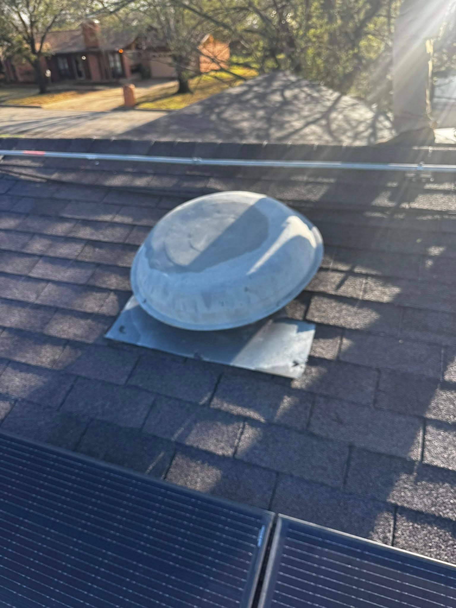 Gray rooftop vent dome on shingles with solar panels and trees in the background