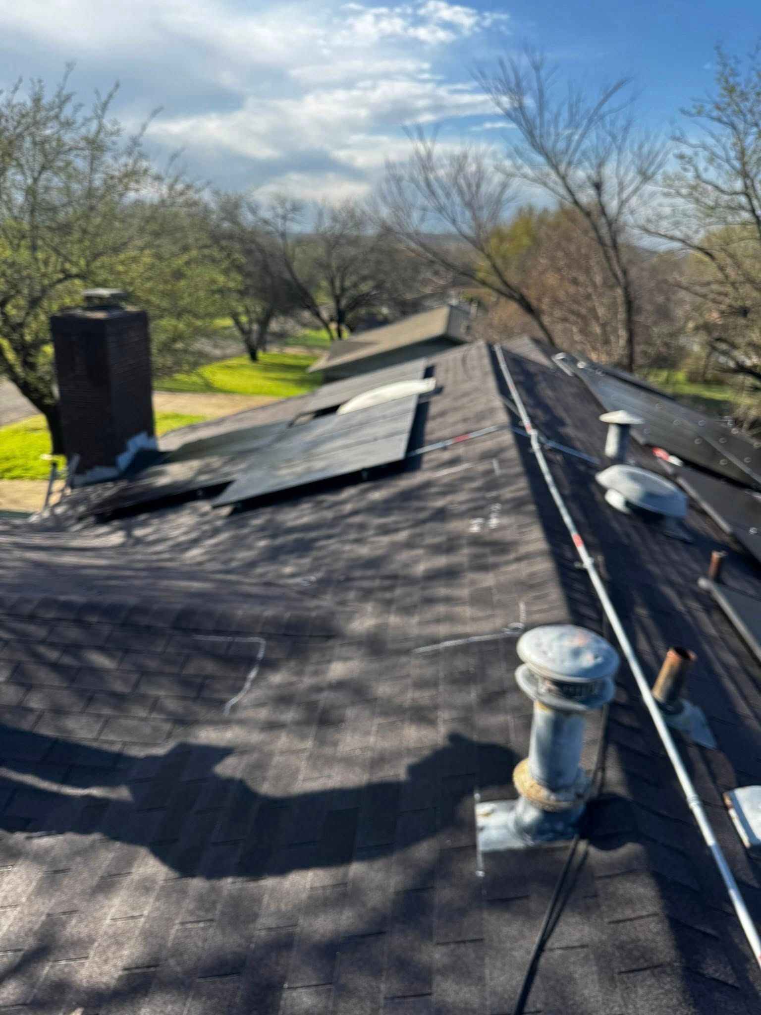 Rooftop with black shingles and vents, viewed along a ridge with trees and blue sky in the background