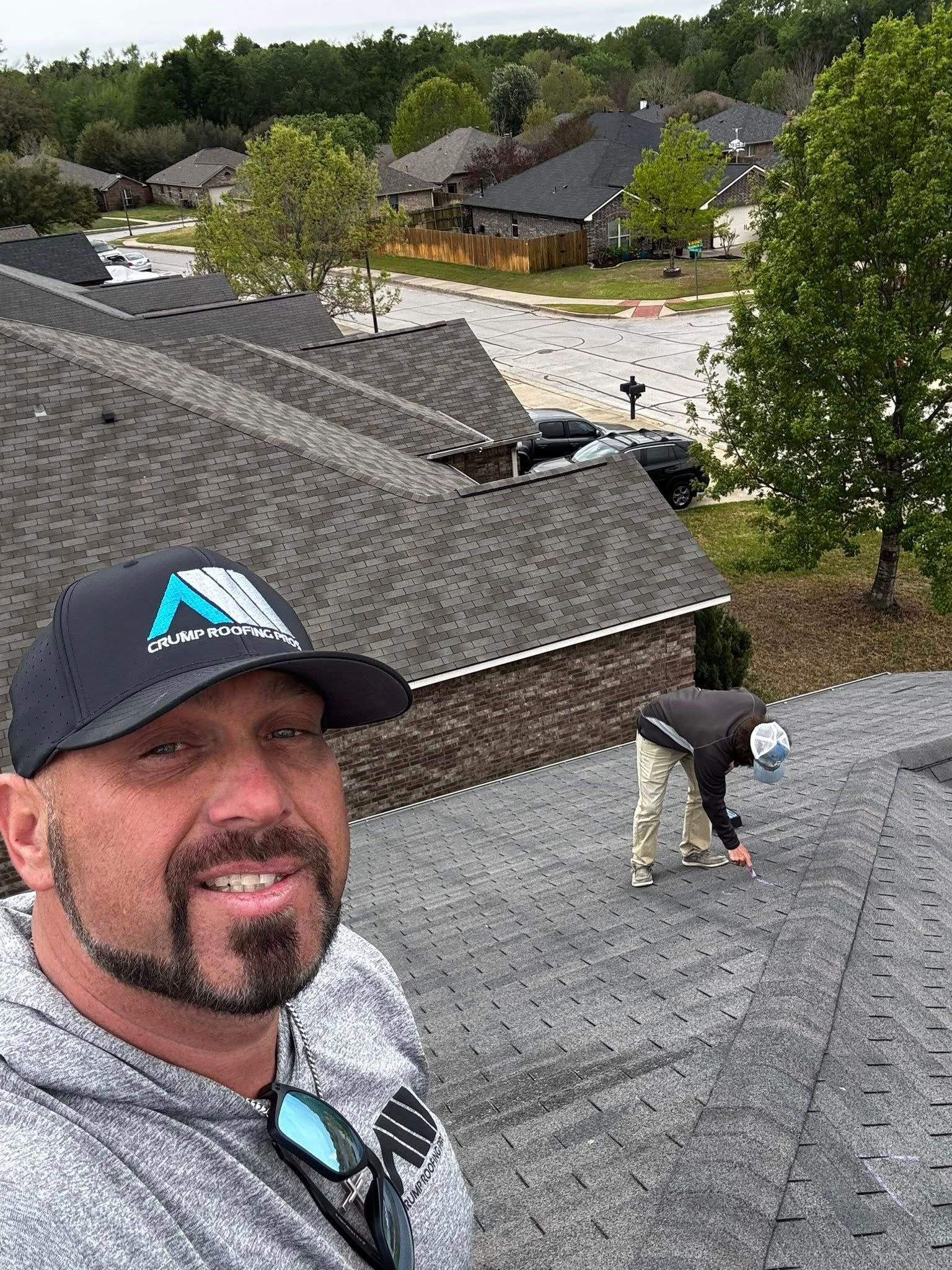 Man in black cap smiling on a shingled roof, with another person working nearby and trees in the background