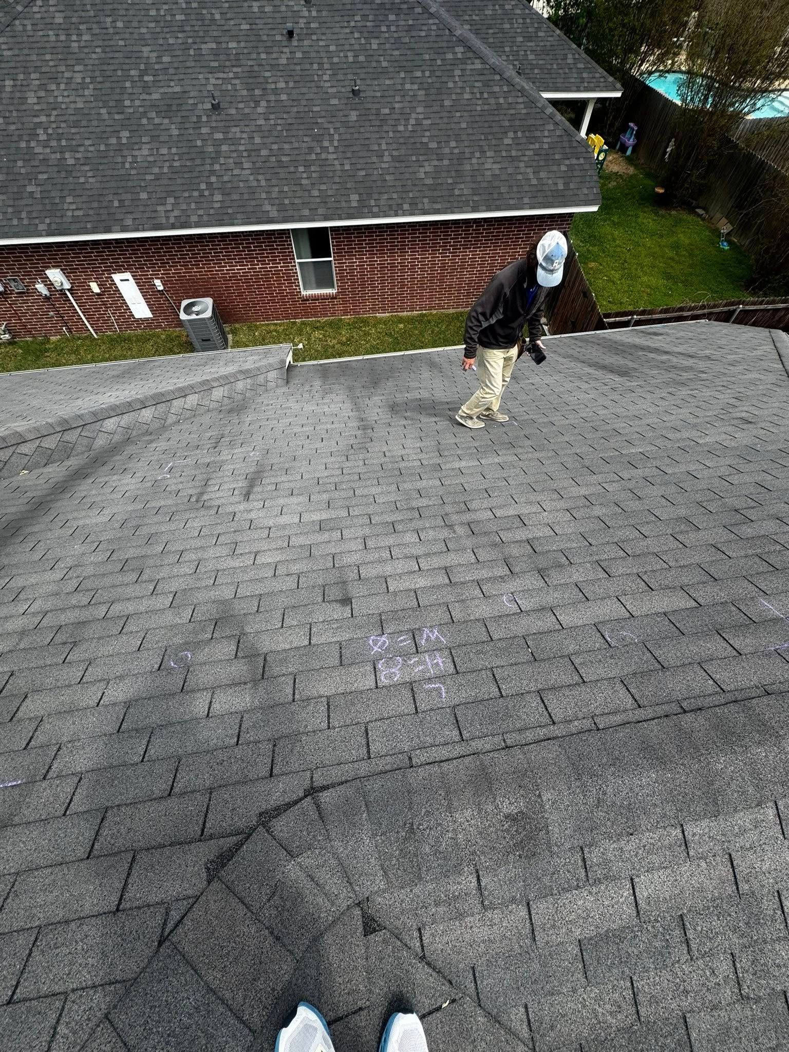 Person standing on a steep shingled roof beside a brick house, holding an object near a vent.