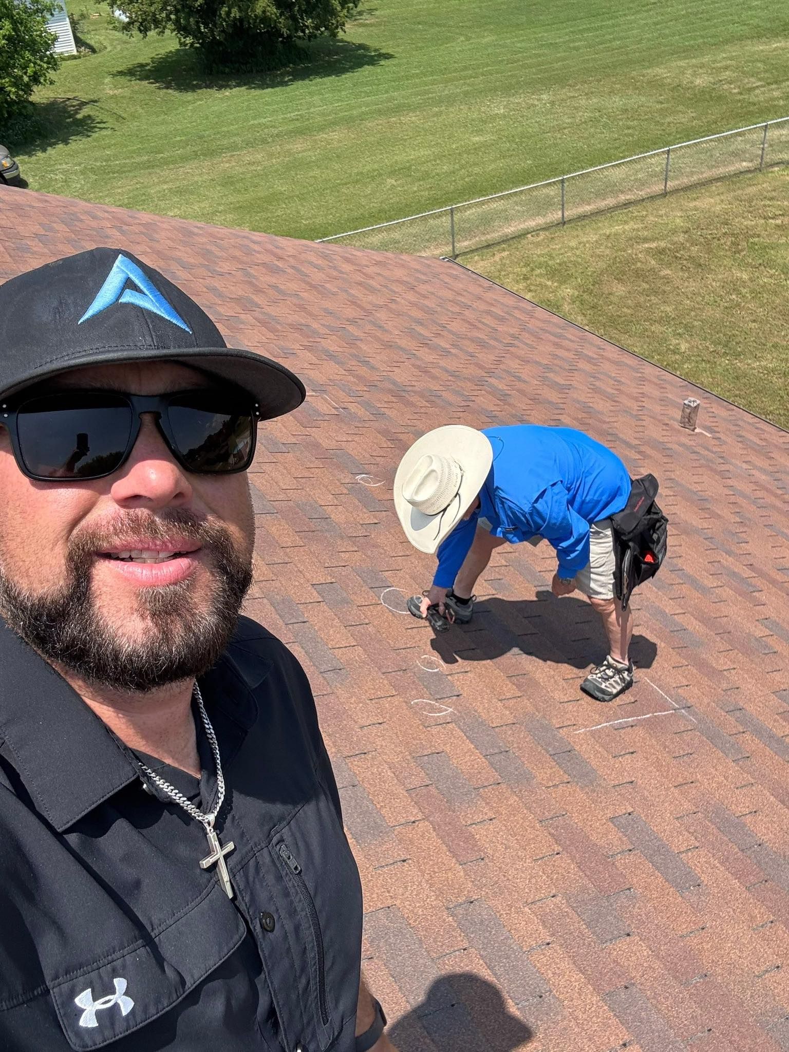Selfie on a rooftop; another person in a blue shirt and sun hat works on the shingles nearby.