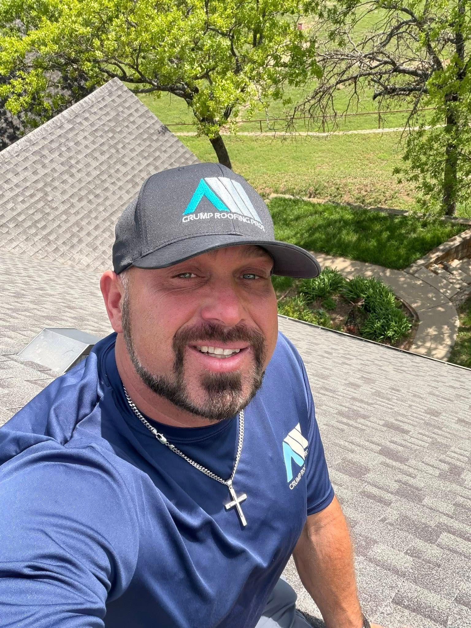 Smiling man in navy shirt and cap taking a selfie outdoors by a tree and fence
