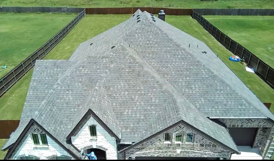 Dark shingled roof over beige brick house corner under a blue sky