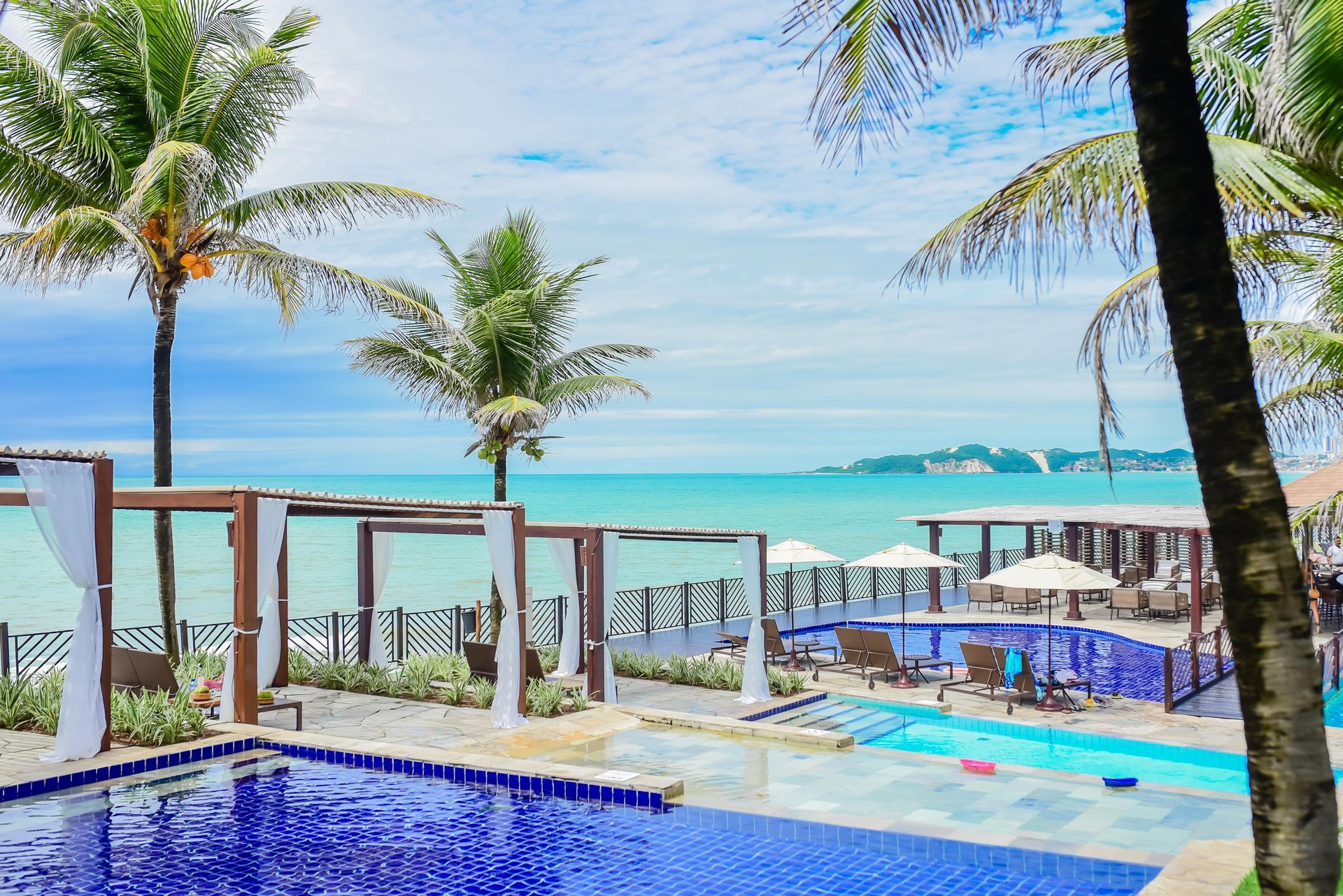 Piscina de resort à beira-mar com palmeiras, água azul e espreguiçadeiras com vista para o mar.