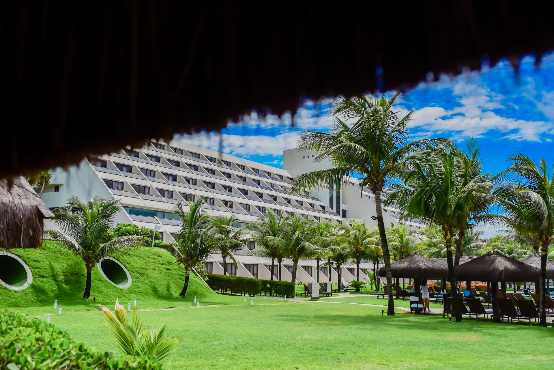 Resort tropical com hotel branco, palmeiras, gramado verde e céu azul com nuvens esparsas.