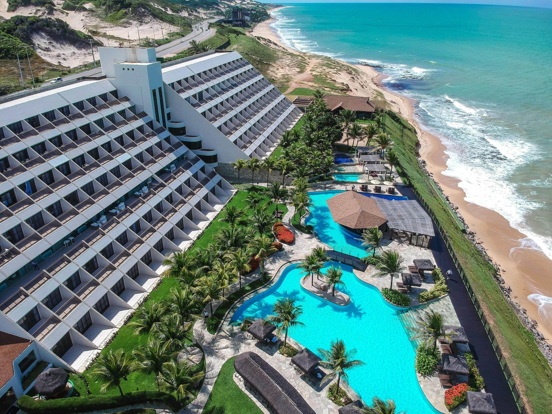 Vista aérea de um resort à beira-mar com piscinas turquesa, palmeiras e ondas do oceano.