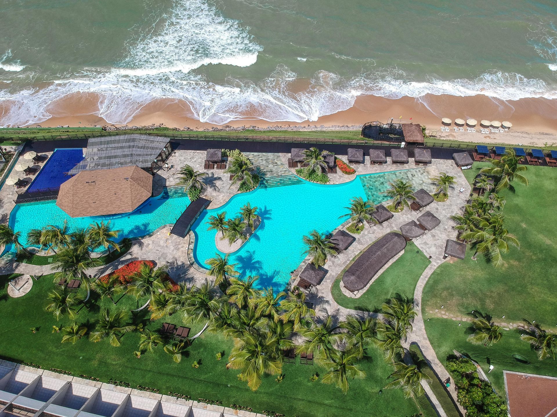 Vista aérea de um resort tropical com piscina turquesa, palmeiras e uma praia com ondas ao fundo.