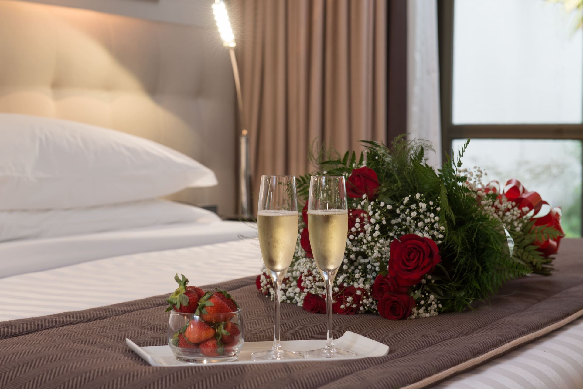 Champanhe e morangos em uma cama de hotel com rosas vermelhas e um arranjo romântico em bandeja.