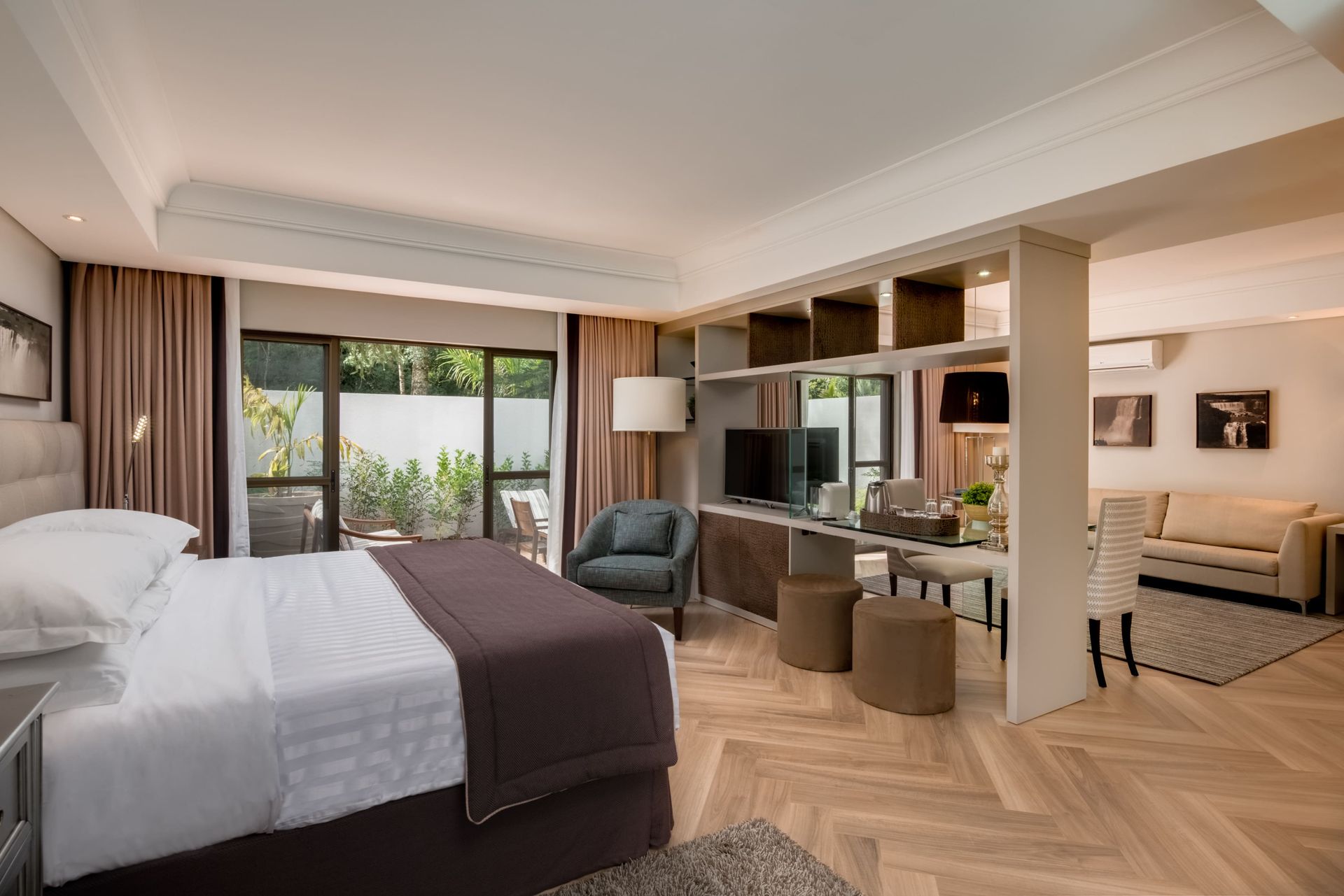 Quarto de hotel moderno com cama king-size, poltronas e portas para varanda, em tons neutros e aconchegantes.