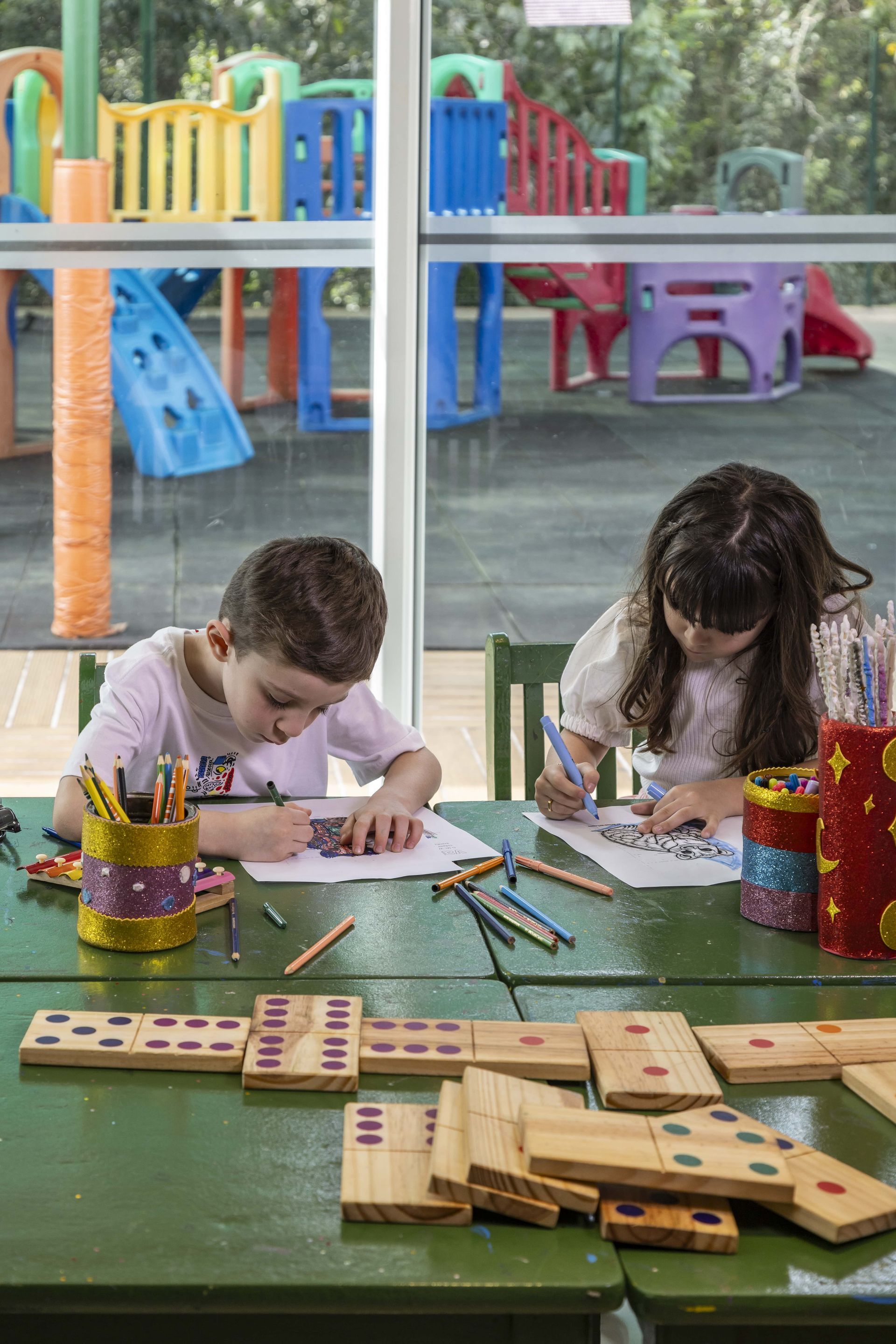 Duas crianças desenhando em uma mesa em um parque infantil colorido.