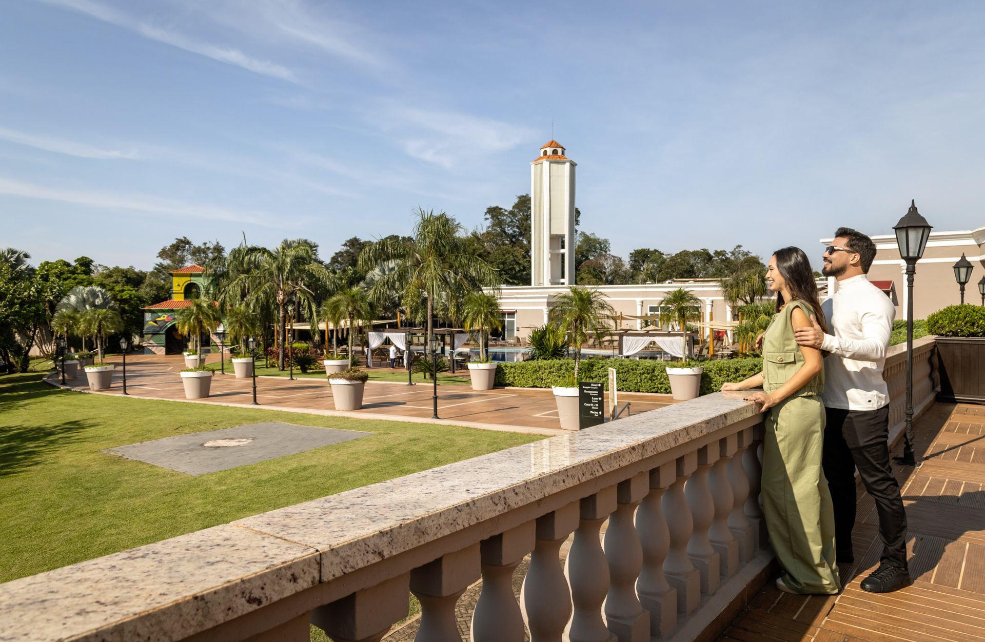 Casal com vista para um jardim paisagístico e uma torre alta em um terraço ensolarado.