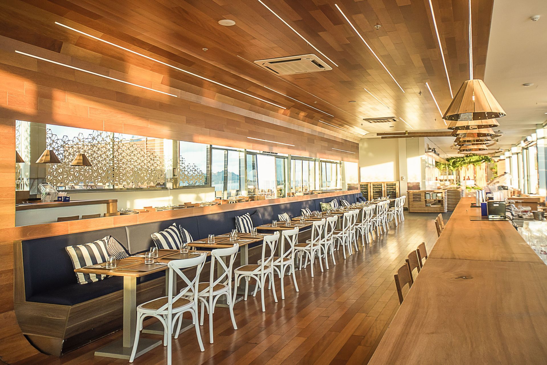 Área de jantar vazia de um restaurante moderno com teto de madeira, balcão extenso e fileiras de cadeiras brancas.