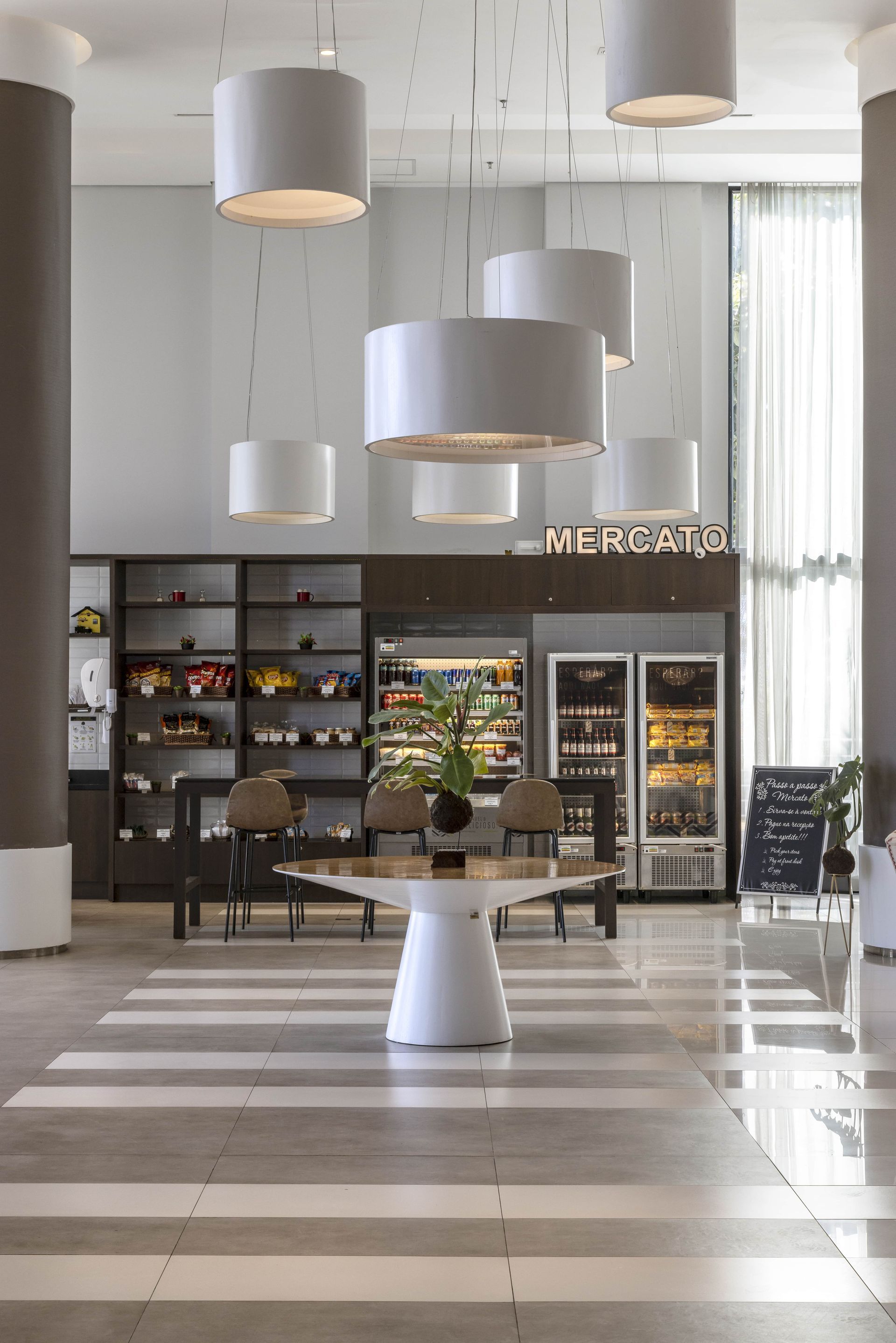 Hall de entrada de hotel moderno com mesa central, piso listrado, luminárias pendentes e uma pequena área de café ou mercado.