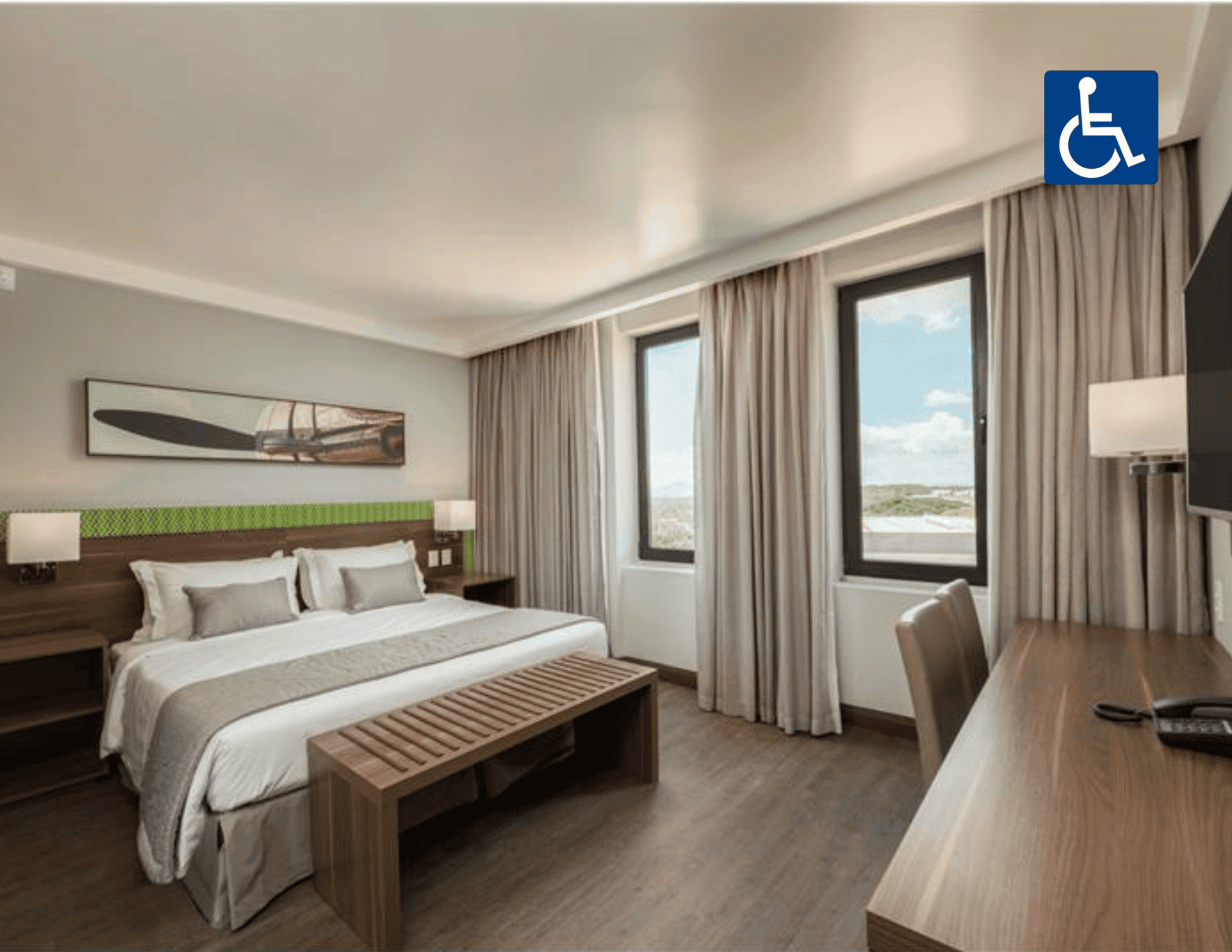 Quarto de hotel acessível com cama, escrivaninha, janelas amplas e placa indicativa de acessibilidade para cadeirantes.