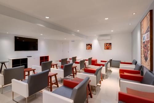 Sala de estar moderna com fileiras de cadeiras vermelhas e cinzas, TV fixada na parede e iluminação embutida no teto.