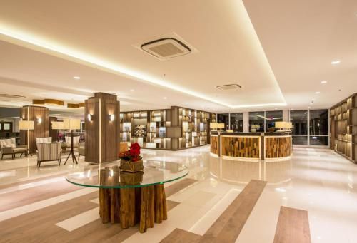 Hall de entrada luminoso do hotel com piso de mármore, mesas de vidro, iluminação aconchegante e área de recepção ao fundo.