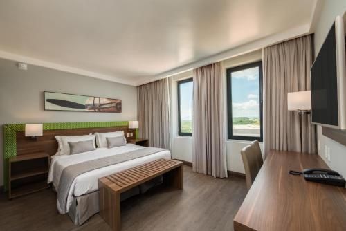 Quarto de hotel moderno com cama king-size, banco, escrivaninha e janelas amplas com cortinas.